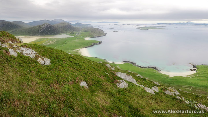 Photo: a grassy foreground interspersed with light grey rocks slopes from left to right. Far below, green grassy small hills and small white sandy beaches. To the left background, a larger beach and small mountains. Islands speckle the sea towards the horizon. The sky is a cloudy grey.