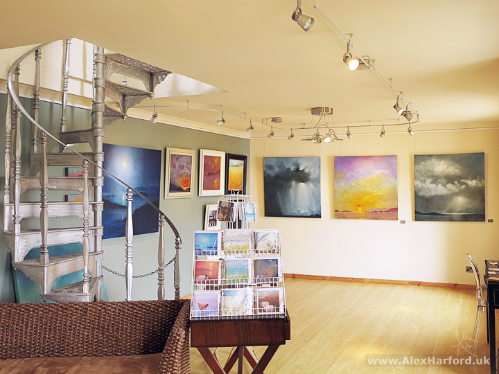 Photo of a small gallery room with greetings cards in the foreground and sky and seascapes on the walls. To the left, a silver spiral staircase.