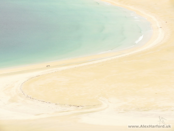 Photo: ariel view zoomed into a beached shaped like a backwards S. The beach is pastel yellow and the see pastel cyan. Small figures walk along the beach to the left.