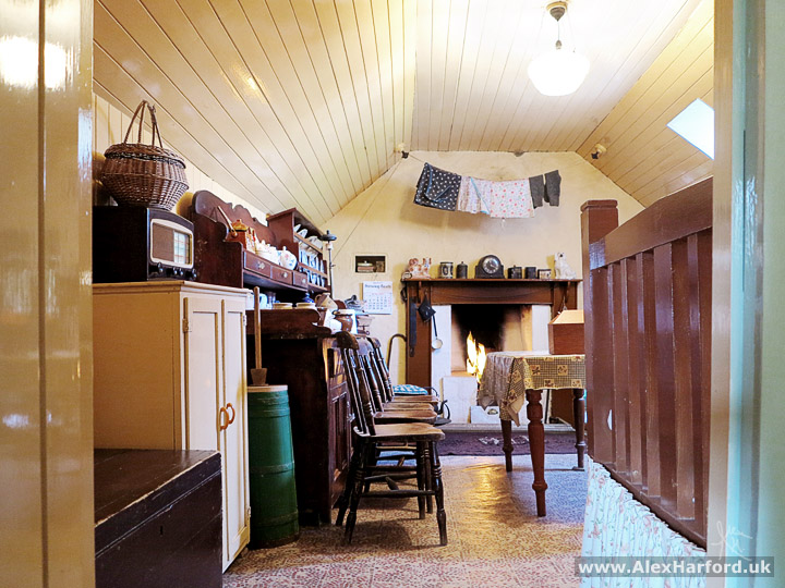 Photo: looking from behind the doorframe, a row of wooden chairs beside a table with a table cloth. A wicker basket on top of an old radio to the left and in the background, washing drying over a fireplace with a clock and ornaments on the hearth. The dominant colours are brown wood and yellow pastel on the walls and ceiling.