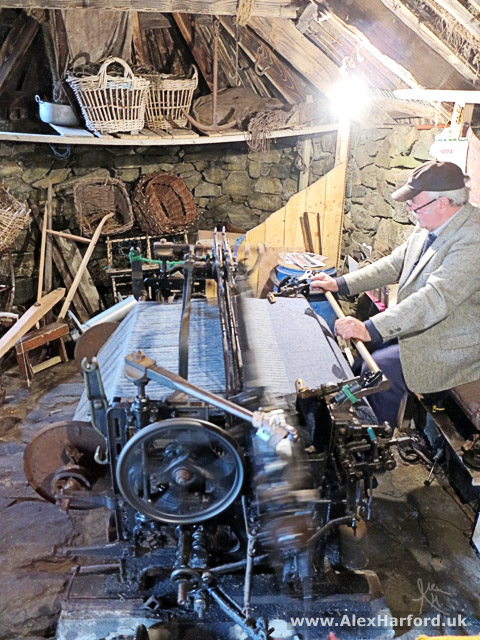 Photo: a man wearing a brown cap sits to the right and works the large loom. In the foreground, the blur of its wheel shows motion. In the background, wicker baskets against a drystone wall.