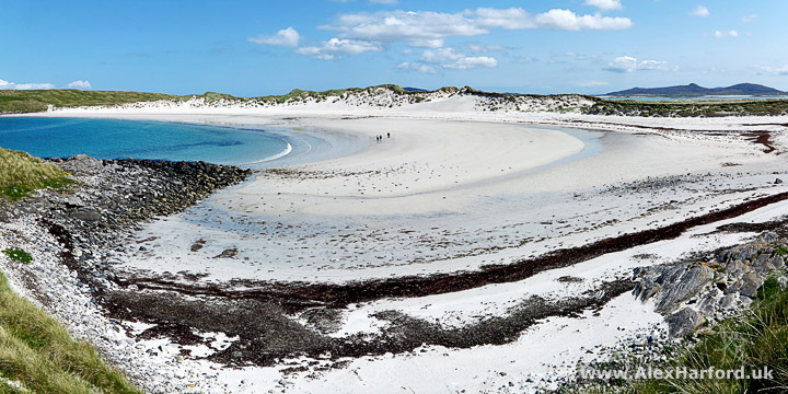 Photo showing a large expanse of white sand curcing around a deep blue sea and backed by sand dunes topped by green grass. On the horizon to the right, a small mountain range. The sky is a deep blue with a few fluffy white clouds.