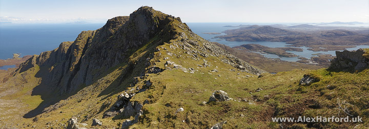 Panoramic photo: to the left a mountain ridge leads to a peak and then down to the dark blue sea. To the right, a grassy foreground with small brown mountains interspersed with lochs. The sky is clear and blue.
