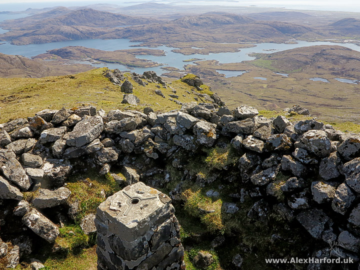 Photo: just off centre in the foreground, a brick trig point. The trig point is surrounded by a lichen and moss-covered wall where many rocks have fallen. Below in the distance, low brown mountains interspersed with long blue lochs.