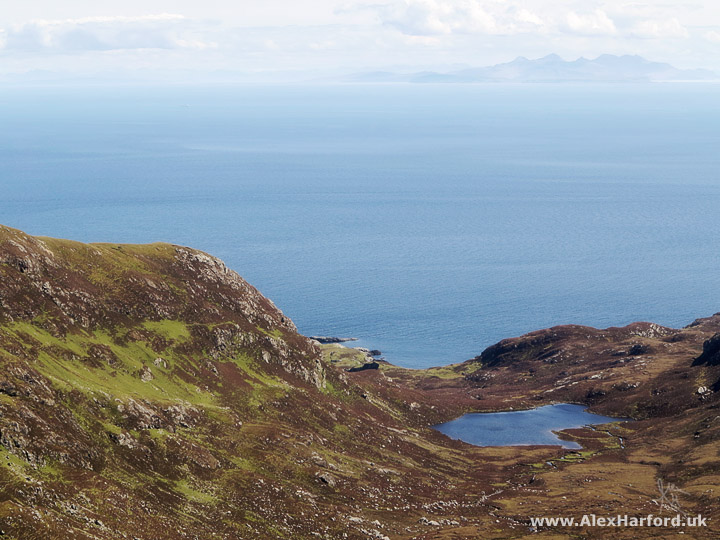 Photo: in the foreground, a brown and green mountain valley slopes downwards towards a loch before a large expanse of blue sea. The faint silouette of a mountainous island appears on the horizon to the right. The sky is light blue with fluffy clouds.